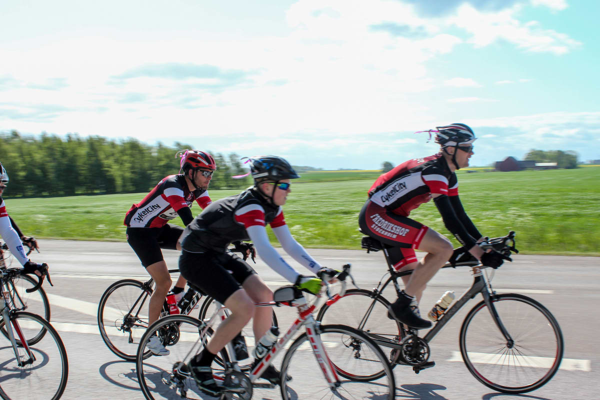Bilden visar fem personer iklädda cykelkläder och hjälmar, cyklandes på en väg under dagtid. I bakgrunden syns ett grönt fält, träd och en ljus himmel med moln. Två av cyklisterna har rosa dekorationer fästa på hjälmarna.