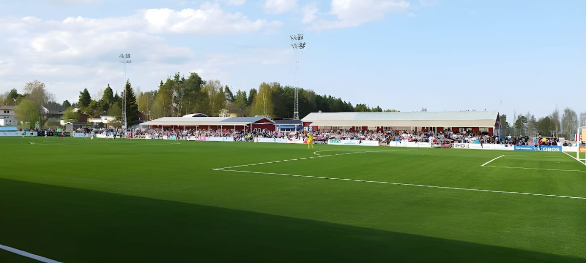 En utomhusbild tagen från en fotbollsplan med konstgräs, omgiven av åskådarläktare och byggnader. I bakgrunden syns skog och en ljusblå himmel med spridda moln. Det är dagtid och solen skiner, vilket skapar skuggor på planen.