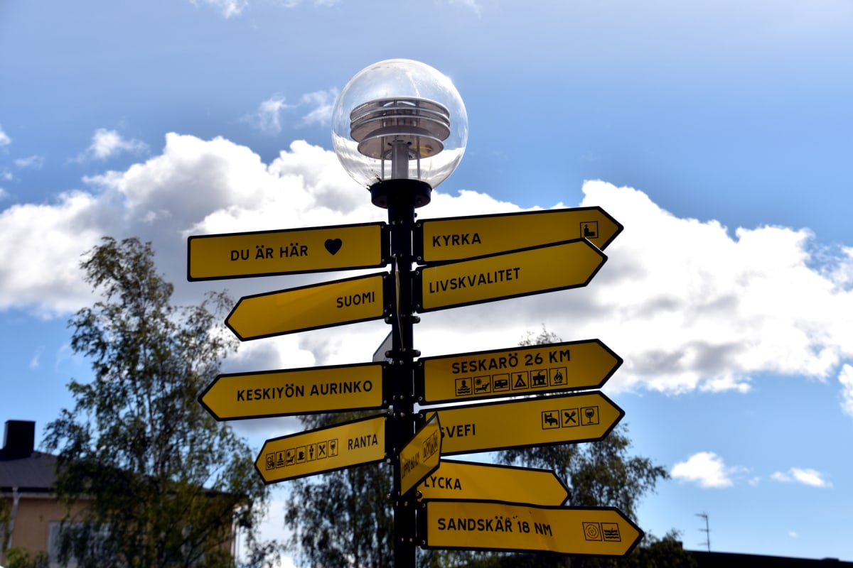 En gulbrun vägvisare med svarta textpilar står mot en blå himmel med vita moln och lövträd i bakgrunden. Pålverkets topp har en rund, transparent armatur med lamphållare som reflekterar ljus.