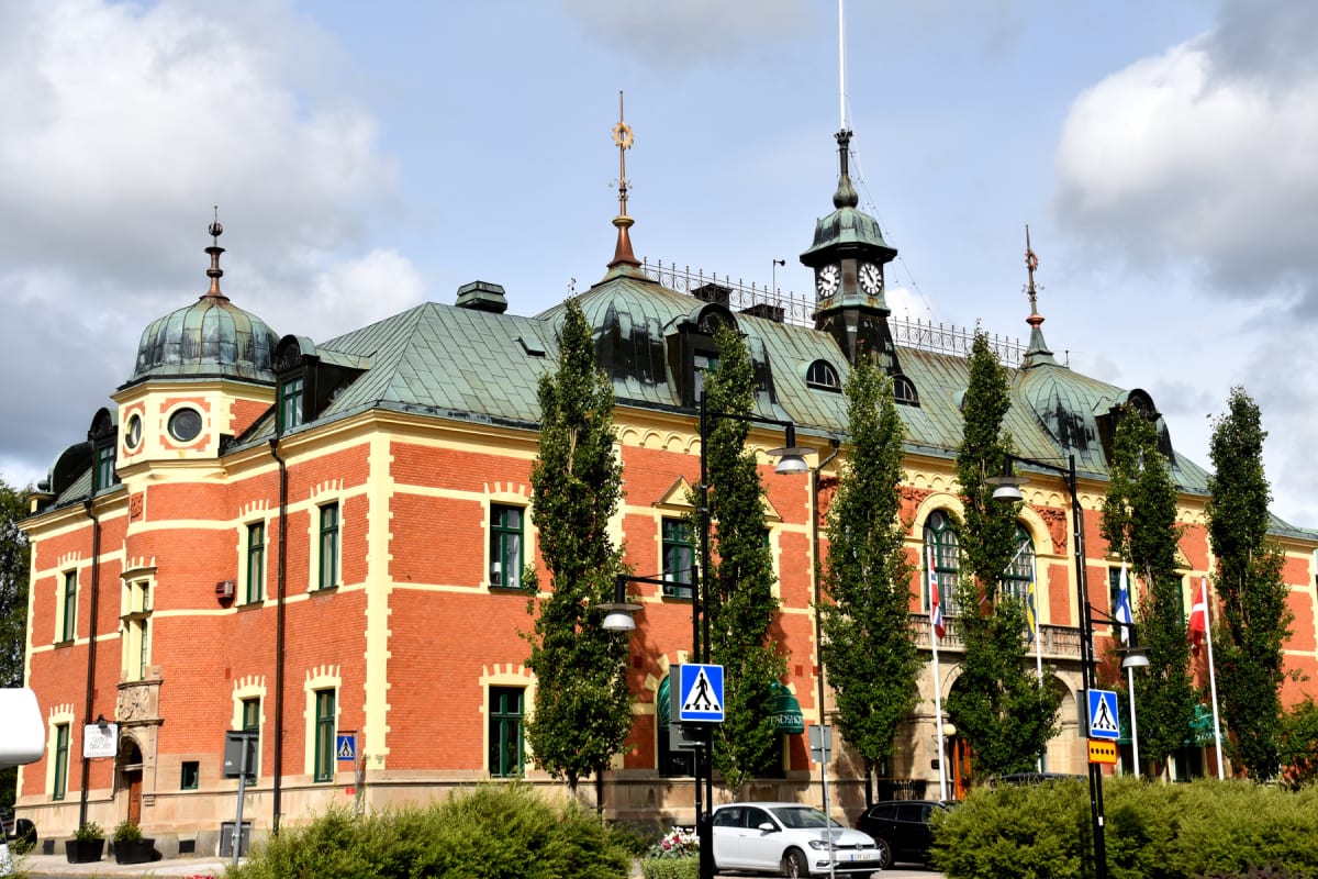 En stor, röd tegelbyggnad med gula detaljer och grönt tak. Flera höga, smala träd står framför byggnaden. På taket syns en klocka i ett torn och andra kupolformade strukturer. En vit bil och en svart bil är parkerade, och en fotgängarskylt syns vid t