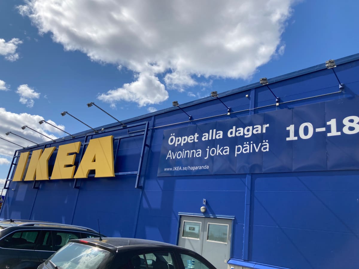 En bild bortom bilarnas tak över en del av en blå IKEA-byggnad med stor gul logotyp och information om öppettider på svenska och finska. En ljusblå himmel med moln syns ovanför byggnaden.