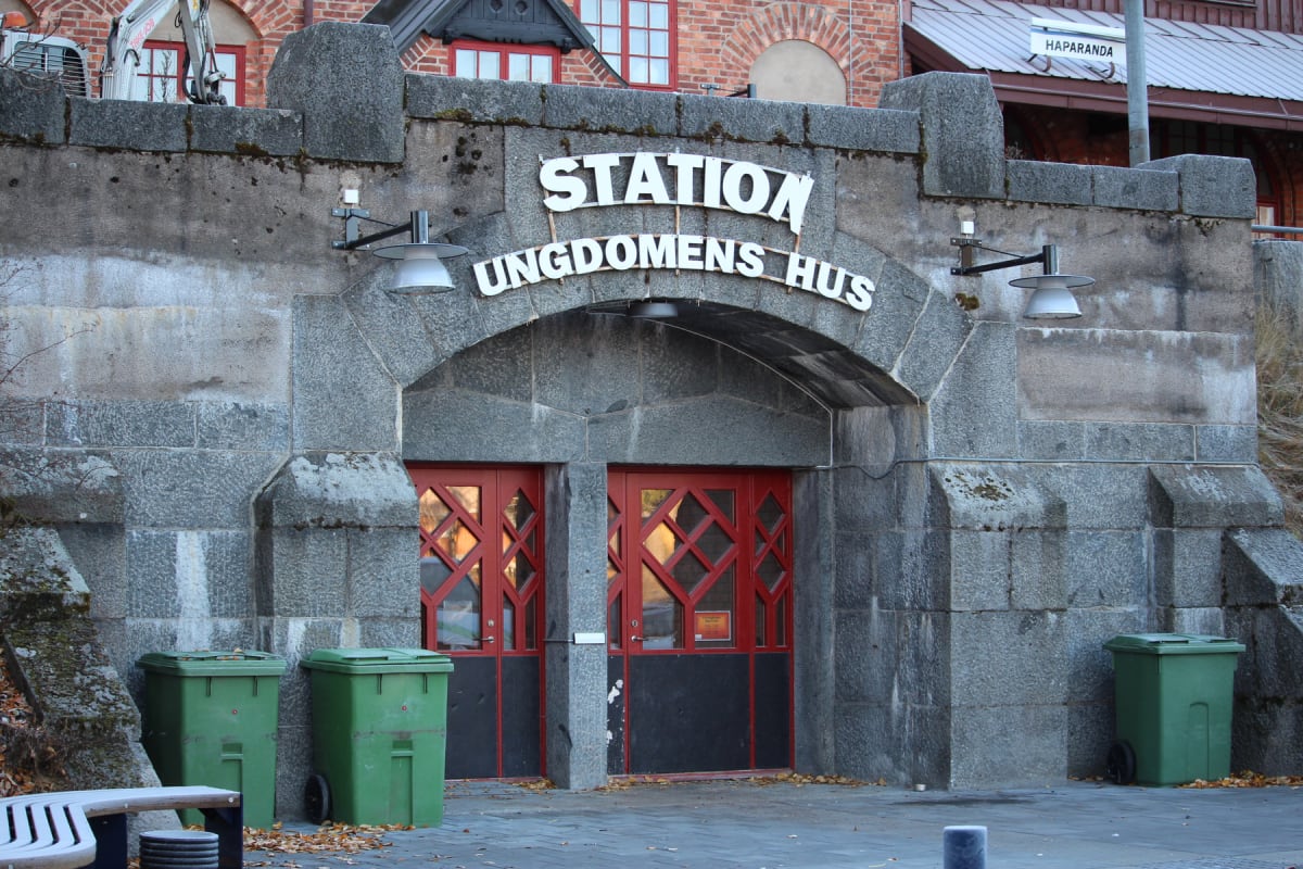 En fasad av grå sten med en välvd ingång som rymmer två röda dörrar med diamantformade fönster. Ovanför ingången står texten "STATION UNGDOMENS HUS". Två gröna soptunnor står utanför dörrarna, och en skylt med texten "HAPARANDA" syns i bakgrunden.