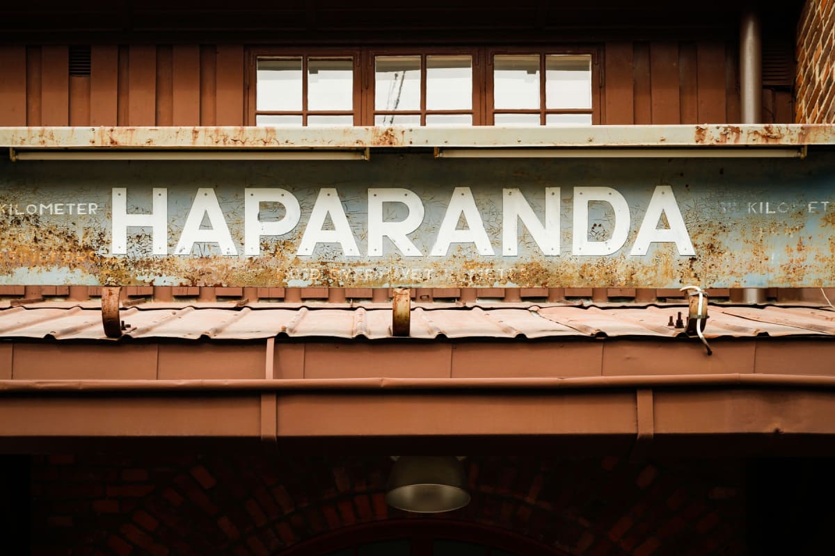 En vertikal bild av en äldre skylt med texten ”HAPARANDA” i stor, vit typografi. Skylten är fäst under ett brunt tak med fönster och ovanför ett mindre brunt tak med en hängande lampa. Skylten är rostig och färgen flagnar, och den sträcker sig från v