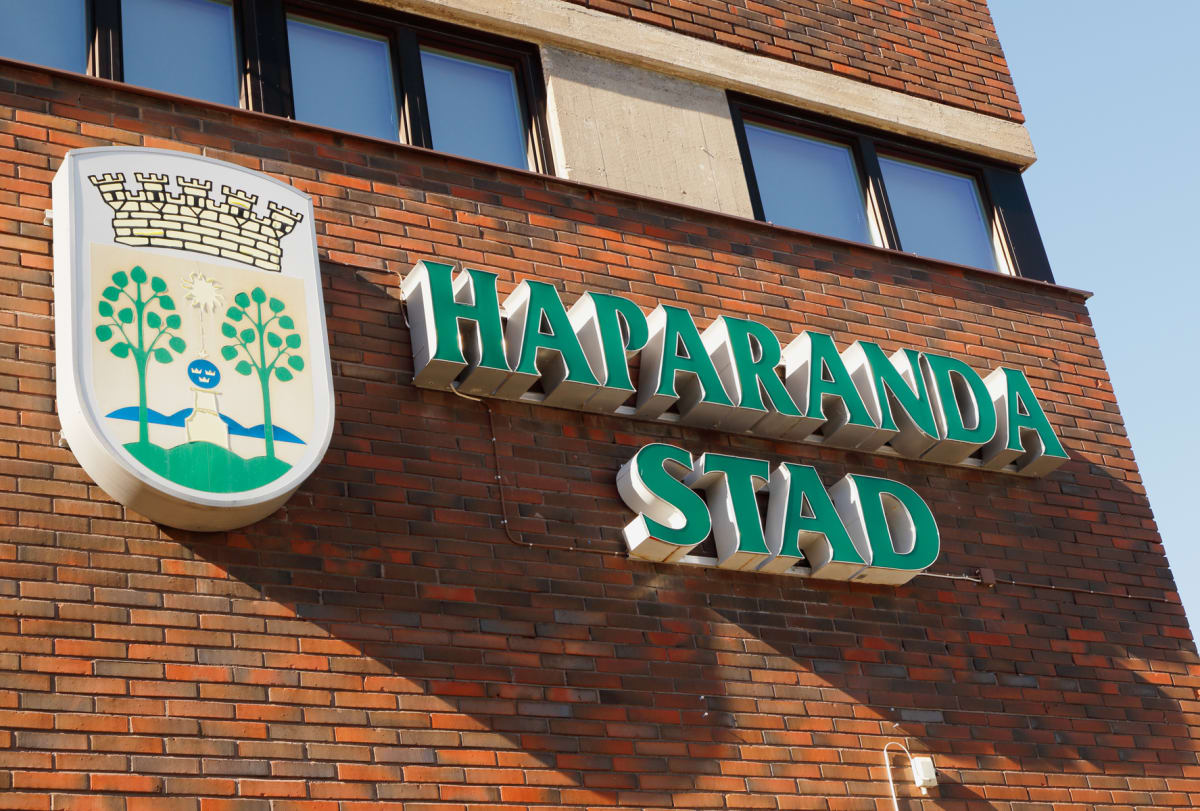 En fasad på en tegelbyggnad med stora skyltar för 'HAPARANDA STAD' och Haparandas kommunvapen intill, under klara blå himlen. Delar av fönster är synliga. Ljuset kommer från sidan, vilket skapar skuggor på fasaden.