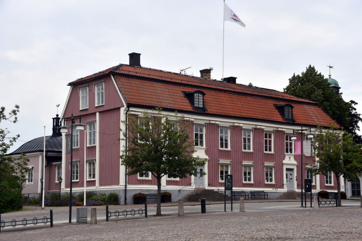 En bild visar en röd träbyggnad med vita fönsterkarmar och en röd tegeltak. En vit flagga med rött märke vajar på en flaggstång på taket. Framför byggnaden finns kullersten, bänkar, träd och cykelställ. En mindre del av en grön kupol kan ses i bakgru