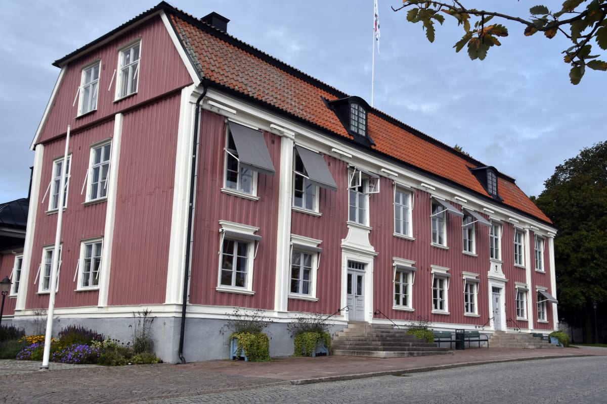 En stor, röd byggnad med vitmålade knutar och fönsterkarmar under en blåsig himmel. Taket är täckt av röda tegelpannor och ett flertal fönster har infällda markiser. Framför byggnaden finns en kullerstensbelagd yta.