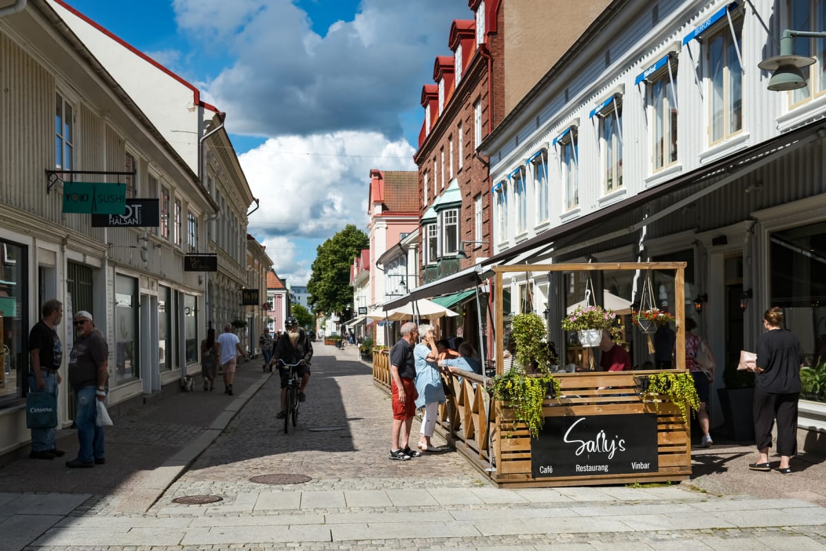En gata kantad av flervåningshus med butiker och restauranger på båda sidor. Människor promenerar längs gatan, och några sitter på en uteservering. En person cyklar i mitten av gatan. Himlen är delvis täckt av moln.
