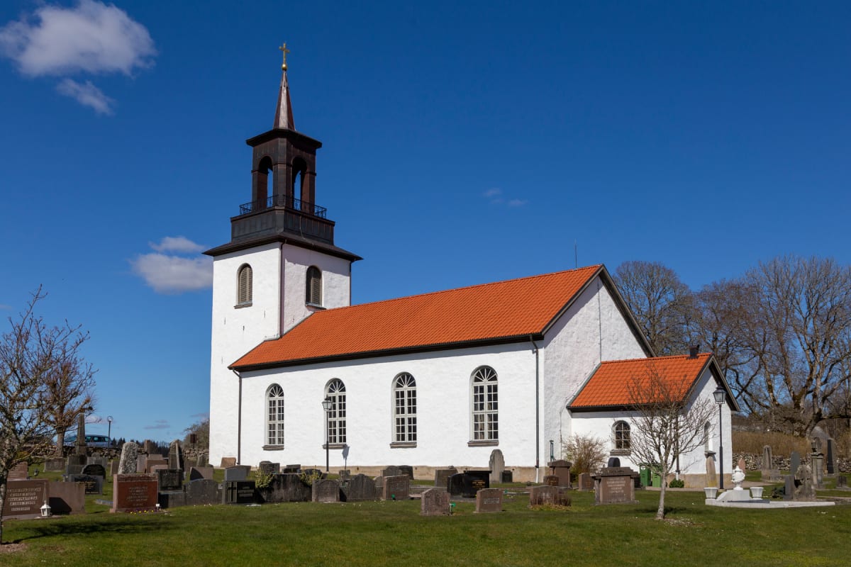 En vit kyrka med rött tak och ett brunt torn med en gyllene toppkors syns under en klarblå himmel. Kyrkogården i förgrunden har flera gravstenar och gräset är grönt, omgivet av kala träd.