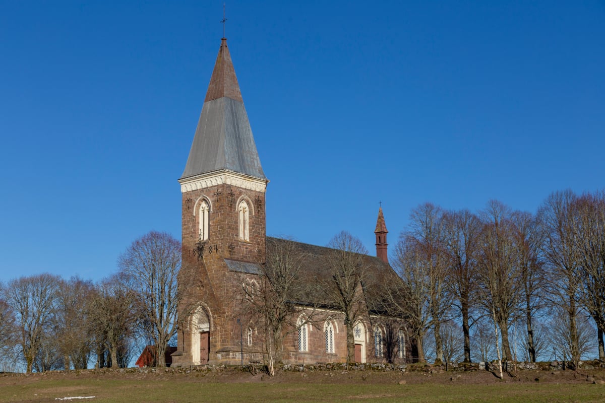 En stor brun kyrka med ett högt, grått spetsigt tak syns i mitten av bilden, omgiven av kala träd. En gräsbeklädd kulle med en stenmur avgränsar kyrkan i förgrunden, och himlen är klart blå.