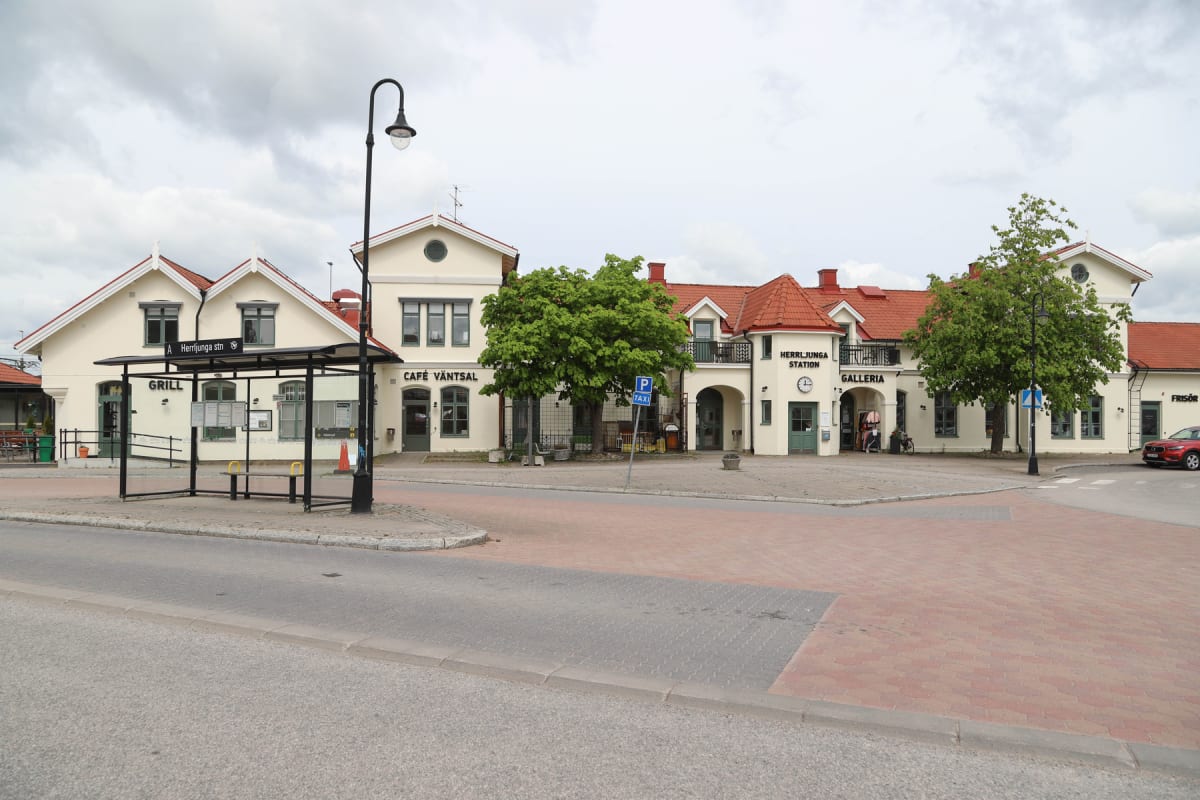 En fasadvy av en gata med en busshållplats i förgrunden. Bakom busshållplatsen syns en beige byggnad med röda tak och flera skyltar med text, inklusive "GRILL", "CAFÉ VÄNTSAL" och "HERRLJUNGA STATION". Träd med gröna löv och en röd bil är också synli