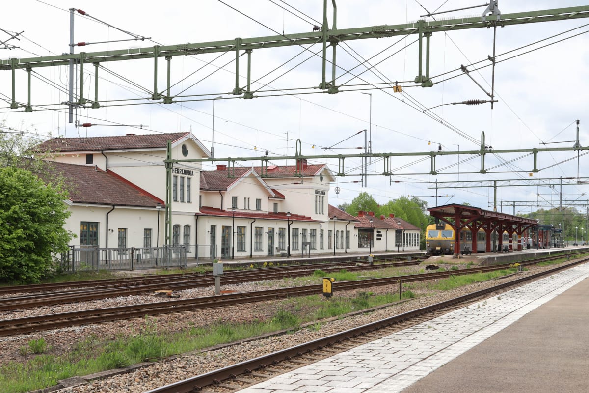 En bild av en tågstation med ett stort stationshus till vänster och tågspår som sträcker sig mot mitten av bilden. Ett tåg står vid en plattform under ett tak på höger sida. Ovanför syns elledningar och elmaster. Stationshuset har ett rött tegeltak o