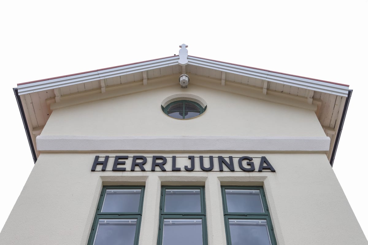 En bild tagen underifrån visar fasaden på en ljus byggnad med mörkgröna fönster. Högst upp sitter en taknock med en liten prydnad ovanför ett runt fönster. Nedanför det runda fönstret står ordet "HERRLJUNGA" med svarta bokstäver. Tre rektangulära fön