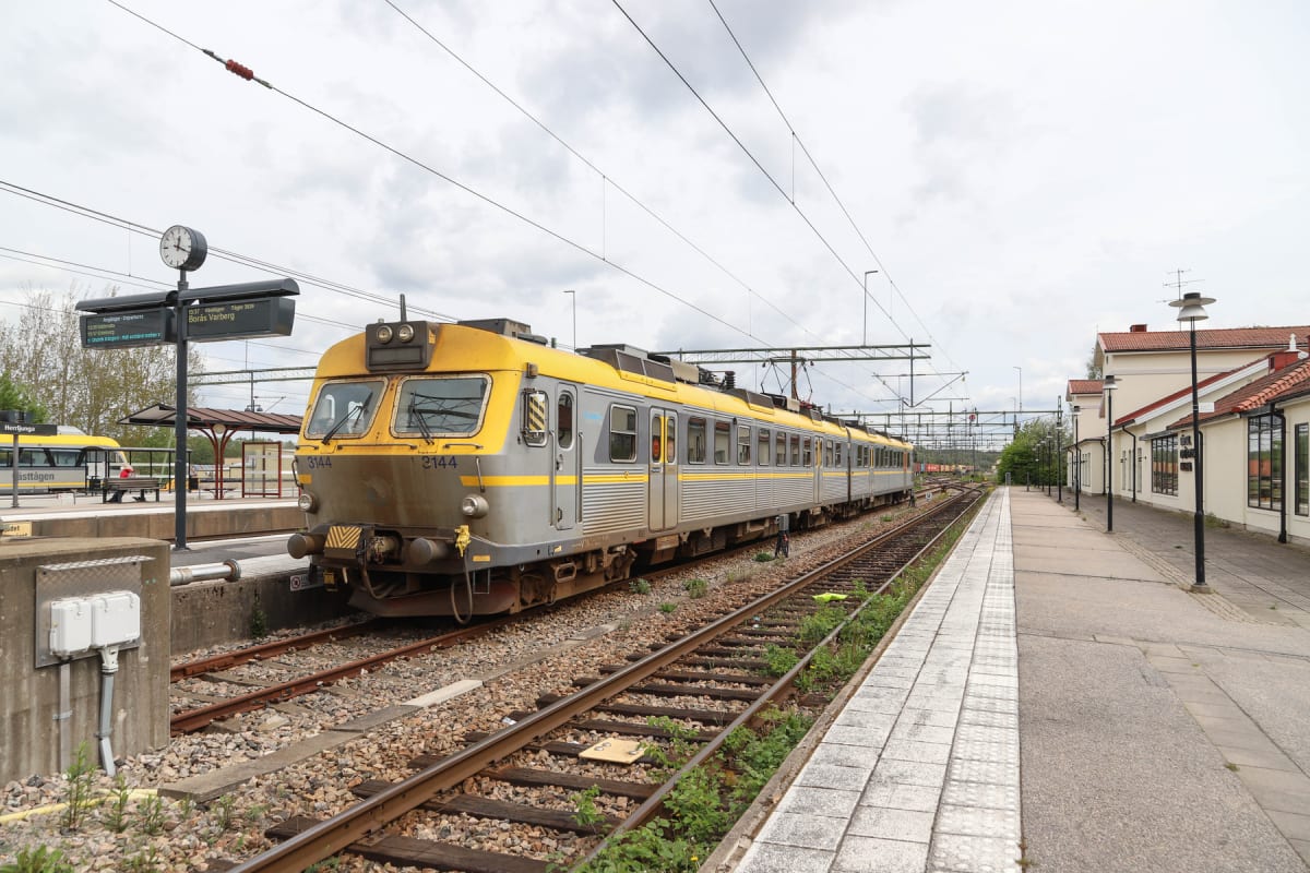 Bilden visar ett tåg med gula och grå vagnar som står vid en perrong på en tågstation under en molnig dag. På vänster sida syns en informationstavla med avgångstider och en klocka. Till höger finns byggnader med röda tak.