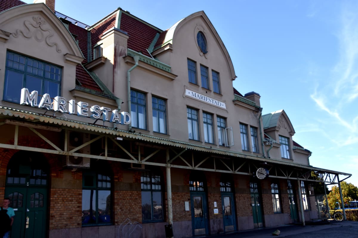 En stor, ljusbeige byggnad med en tegelfasad nedtill och ett rött tegeltak under en klarblå himmel. På fasaden står "MARIESTAD" skrivet med stora bokstäver. Byggnaden har flera fönster, och vissa står öppna. Vid en dörr står en person, och en cykel s