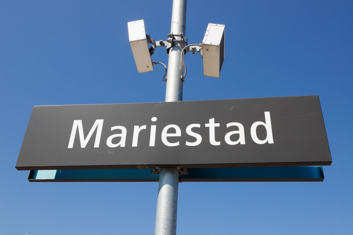 En svart skylt med texten ”Mariestad” i vitt är monterad på en metallstolpe. Ovanför skylten sitter två rektangulära högtalare fästa i stolpen. Allt är placerat mot en klarblå himmel.
