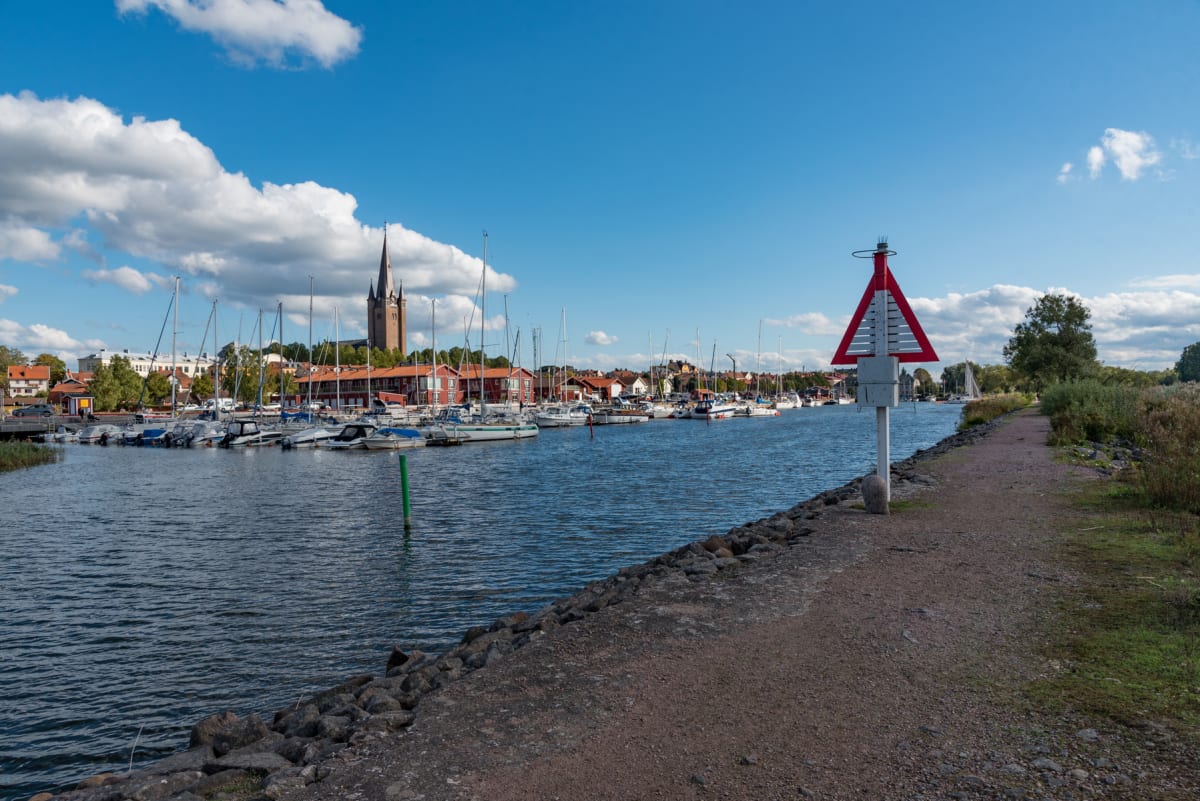 En bild som visar en småbåtshamn med många segelbåtar och motorbåtar. I bakgrunden syns en kyrka med en spetsig tornspira och röda hus. I förgrunden till höger finns en grusväg längs med vattnet och en röd triangelformad sjömärkeslykta.