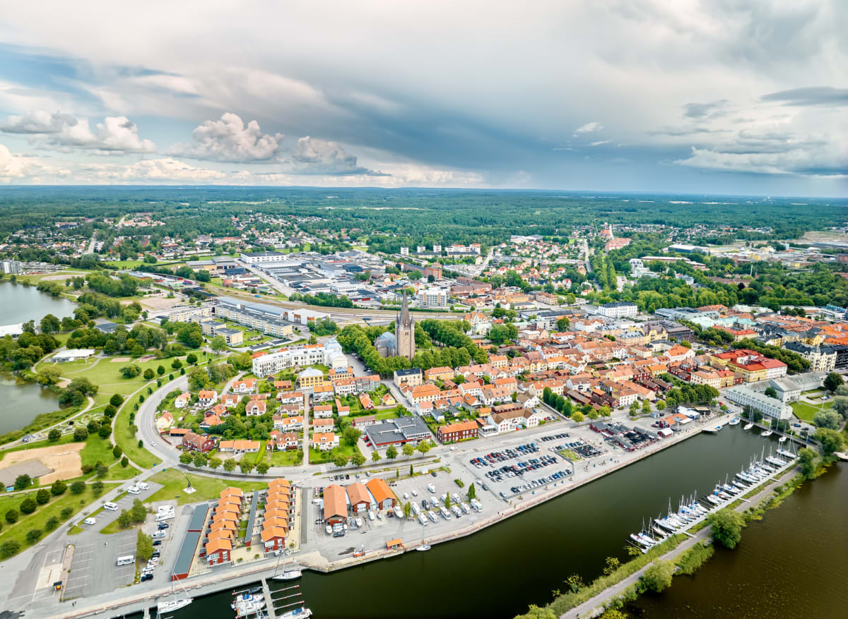 Flygfoto över en stad intill en större vattenyta. Staden består av bostadsområden med röda tak, en kyrka med spira, industriområden, en småbåtshamn med segelbåtar och större grönområden. Himlen är delvis täckt av moln.
