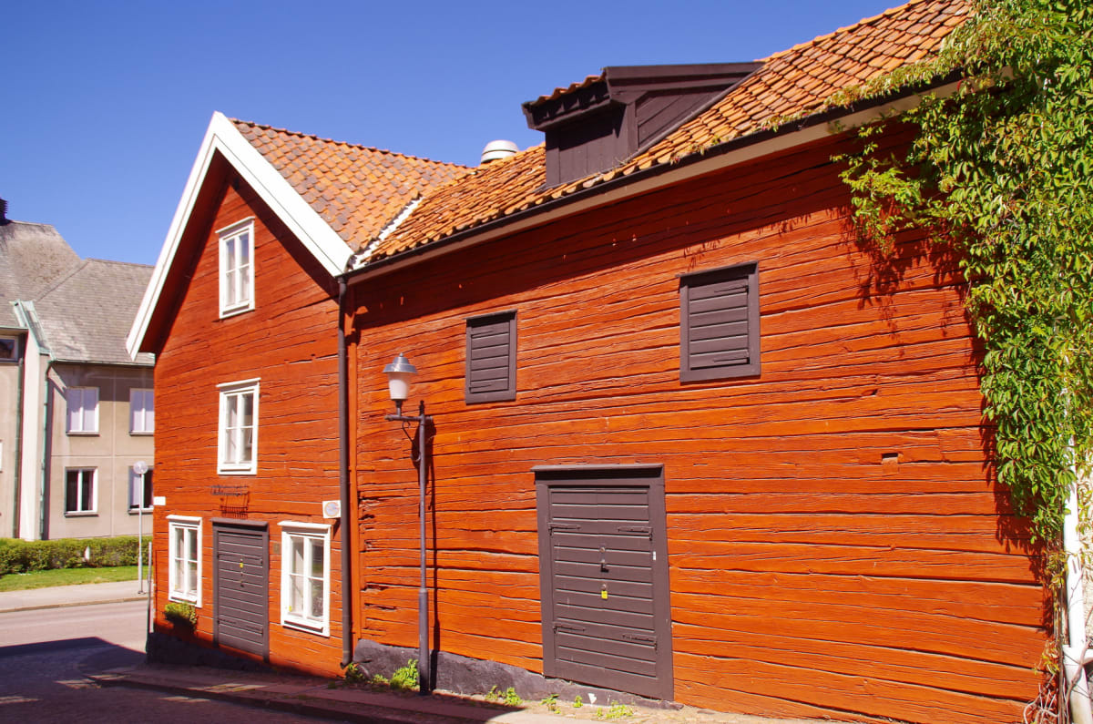 En röd träbyggnad under en klarblå himmel. Väggarna har synliga träplankor, fönster med vita karmar och två dörrar i mörkare brunt. En klätterväxt hänger längs byggnadens högra sida och ett gatlyse står intill fasaden. Taket är täckt med tegelpannor