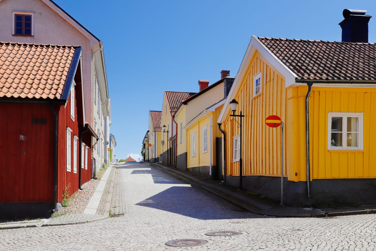 En gata med gatsten och hus i olika färger, främst rött och gult. Gatan lutar uppåt och på husväggarna syns lyktstolpar och en stoppskylt. Himlen är klarblå och skuggor faller över gatstenen.