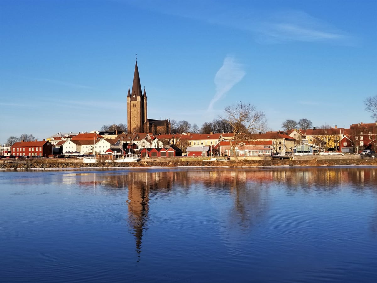 En bild som visar en stadssiluett med en stor stenkyrka med spira i centrum, reflekterad i ett lugnt vatten. Hus med rött tak kantar flodbanken, en del av dem är vita. Träd utan lövverk syns längs horisonten. Himlen är klarblå med några vita moln.