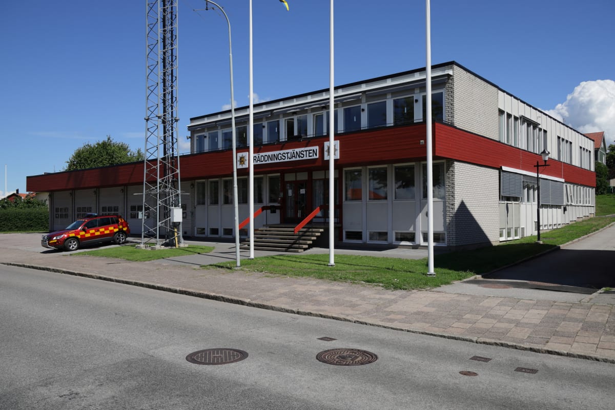 En stor, vit och röd byggnad med texten "RÄDDNINGSTJÄNSTEN" på fasaden, flaggstänger framför, en stor antennmast till vänster och ett rött fordon med gula markeringar parkerat utanför. Himlen är blå med några vita moln. Vägen och trottoaren syns i fö