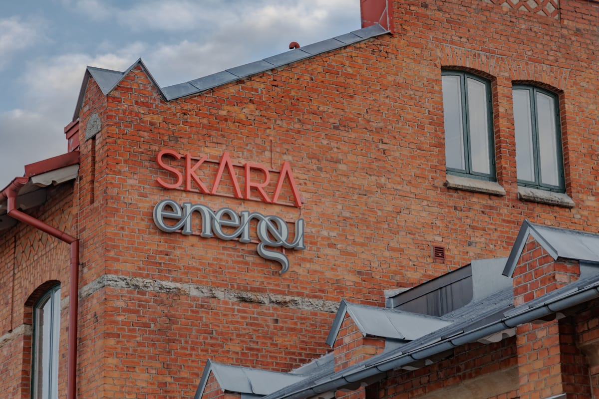 En stor del av en tegelbyggnad syns, med en skylt som lyder "SKARA energi" i rött och grått. Två välvda fönster med mörkgröna bågar är synliga, samt flera taklisterna och en röd stupränna. En blåsig himmel med moln ses i bakgrunden.