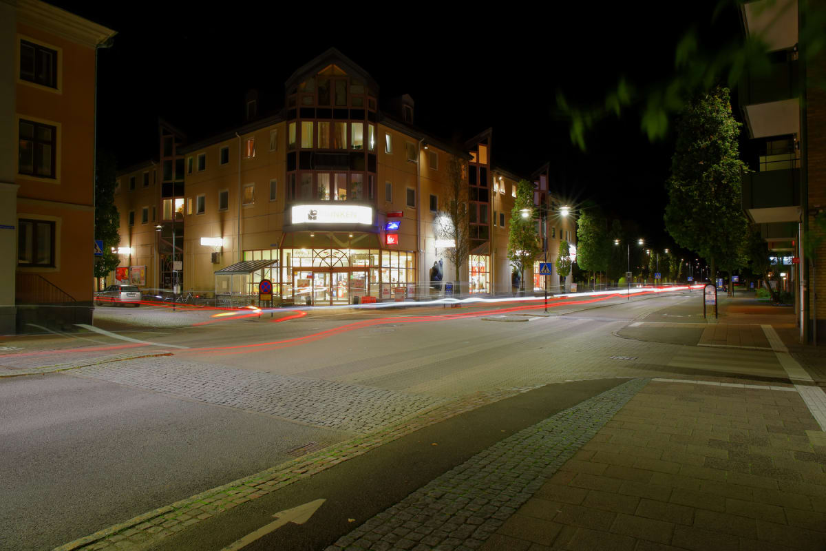En nattbild som visar en gatukorsning med ett flervåningshus upplyst i bakgrunden. Röda och vita ljusspår från fordon syns på vägen, vilket tyder på lång exponering. Träd och ljusstolpar kantar gatan.