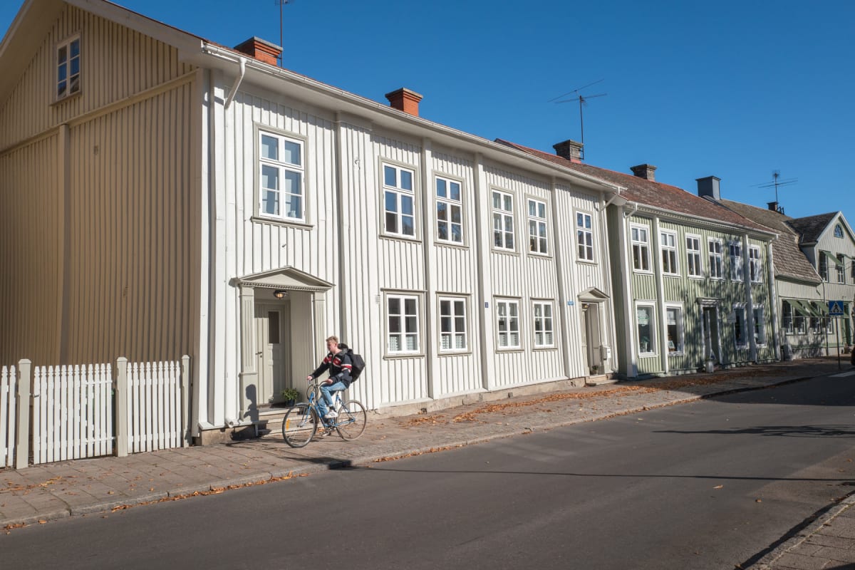 En gata med flertalet träfasadhus, varav de tre första har vita fasader och det fjärde en ljusgrön. En person på cykel syns på trottoaren vid ett av de vita husen. Löv på marken indikerar höst. Byggnaderna har många fönster och skorstenar på taken un