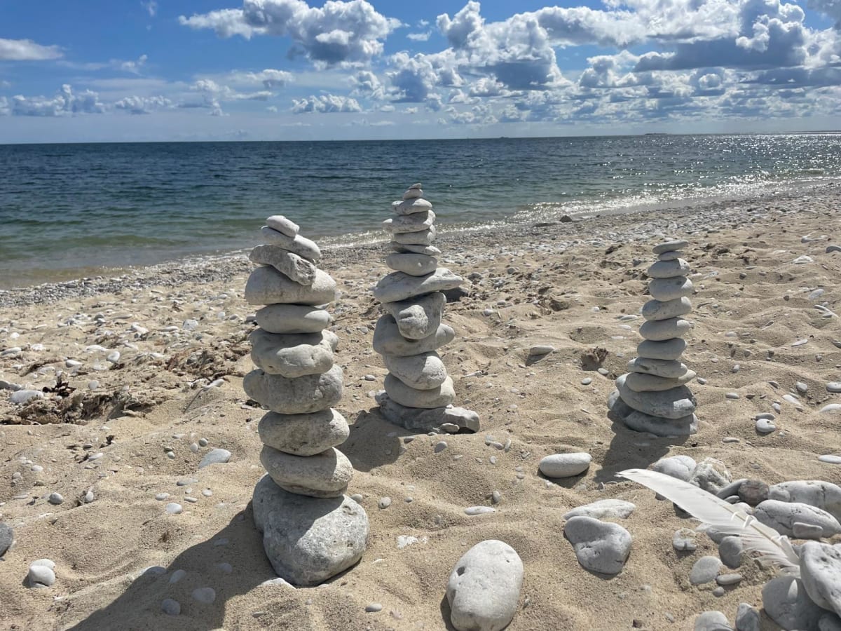 Bilden visar en strand med sand, stenar och vatten i förgrunden. Tre staplar av ljusa, runda stenar står på sanden. I bakgrunden syns ett hav under en molnig blå himmel. En ljus fjäder ligger på sanden till höger.