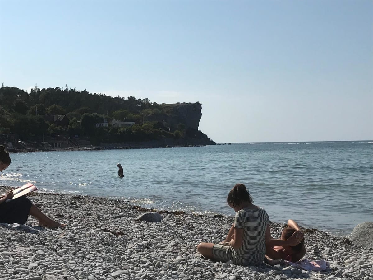 En bild tagen från en stenstrand med en bukt i förgrunden, omgiven av en brant, trädbevuxen kustlinje och en hög, kal klippformation i bakgrunden. Flera personer ses vid vattnet, varav en badar och andra sitter på stranden. Himmel är klarblå.