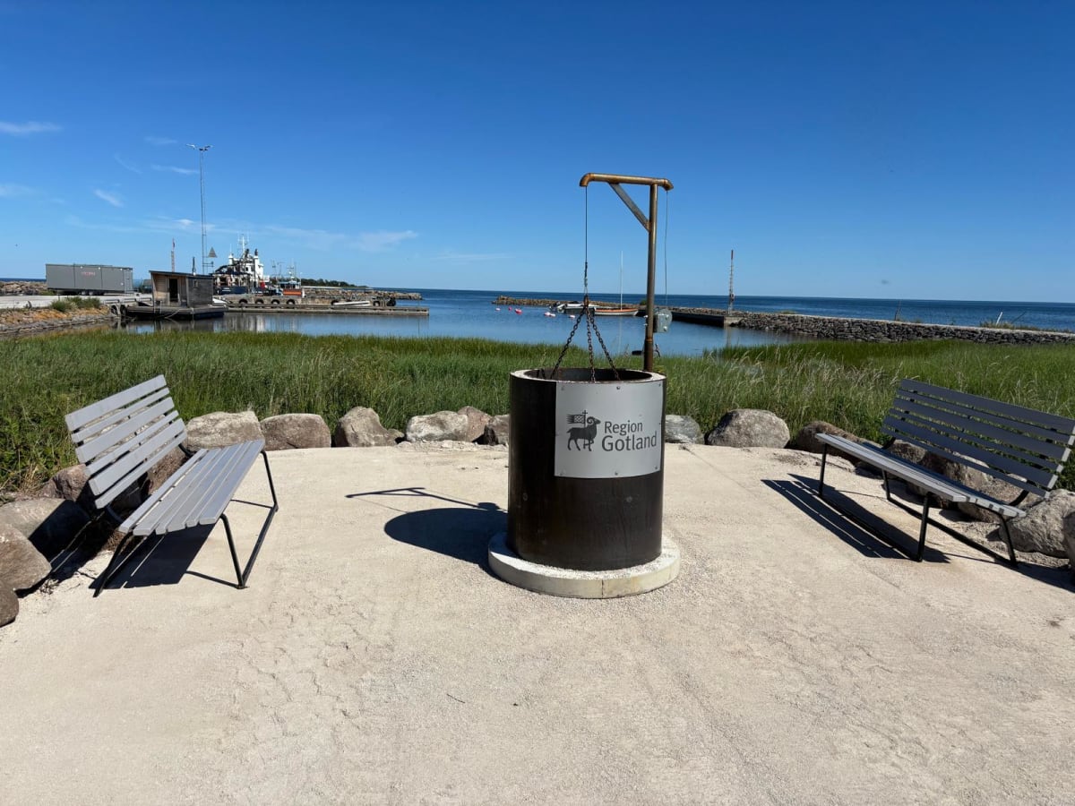 En utomhusbild tagen under dagtid med klarblå himmel, föreställande två bänkar som vetter mot en vattenyta med en brygga och båtar. I förgrunden står en skylt med texten "Region Gotland". Miljön omges av stenar och gräs.