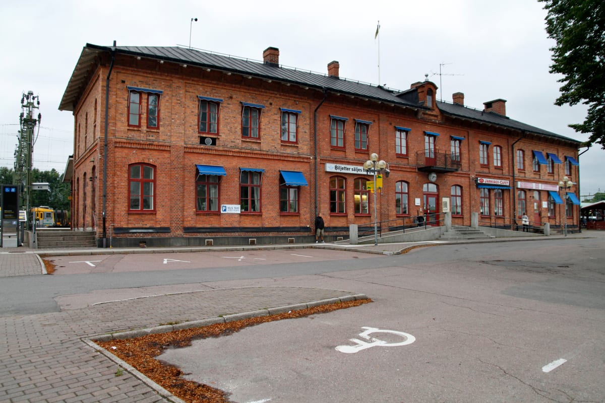 En stor, röd tegelbyggnad med många fönster och blå markiser under en gråhimmel. Byggnaden har ett svart tak med skorstenar. Framför byggnaden finns en asfalterad yta med parkeringsmarkeringar, löv på marken och en gul tågvagn syns i bakgrunden till