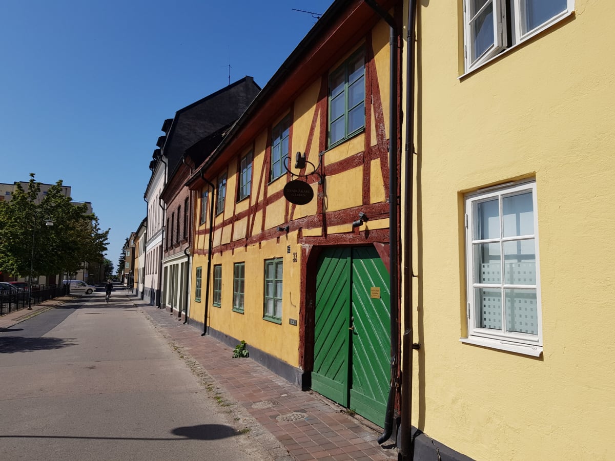 En gata med traditionella byggnader i stadsmiljö under dagtid. Till höger syns en gul putsad byggnad och en korsvirkesbyggnad med gul fasad, röda bjälkar och gröna fönsterluckor/dörrar. En person cyklar borta vid horisonten.