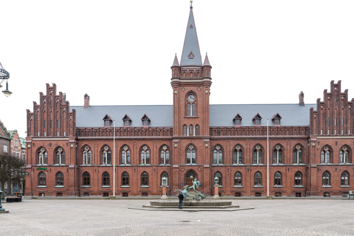 En stor röd tegelbyggnad med detaljerad arkitektur och ett klocktorn, sedd framifrån. Framför byggnaden finns ett torg med gatsten och en fontänprydnad. En person står vänd mot fontänen.
