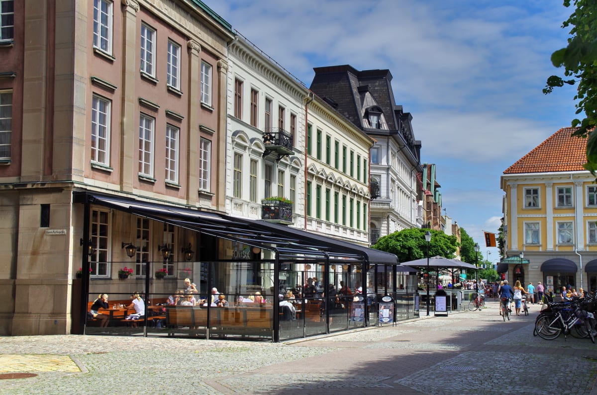 En gata med gatsten i en stad kantad av flervåningshus med restauranger med uteserveringar på en solig dag. Människor syns sitta utomhus vid bord och cykla, samt gående på gatan. Himlen är ljusblå med vita moln.