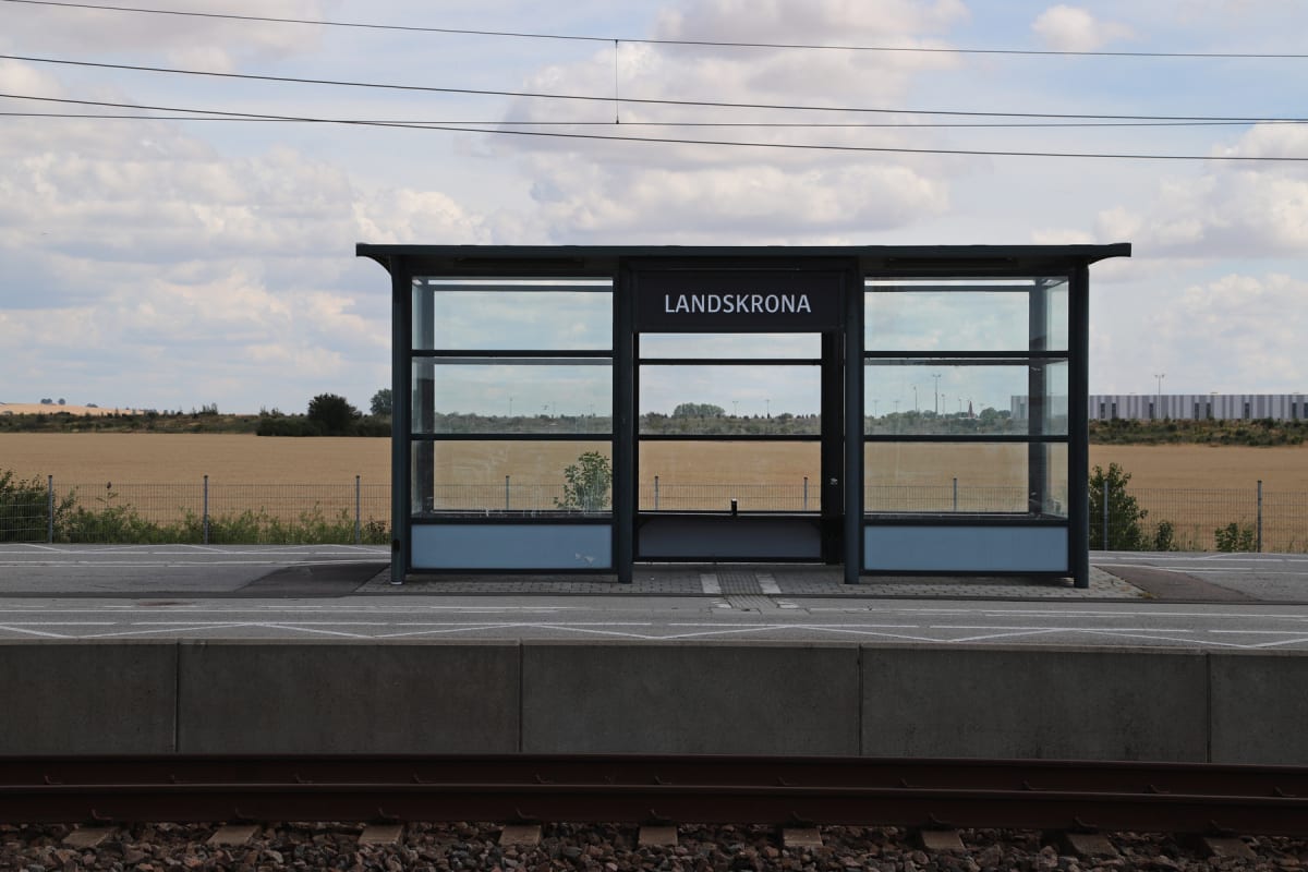 En modern väntkur med texten "LANDSKRONA" står på en tågplattform intill ett järnvägsspår. I bakgrunden syns ett stort fält med en byggnad i horisonten och en molnig himmel ovanför. Flera ledningar sträcker sig över himlen.