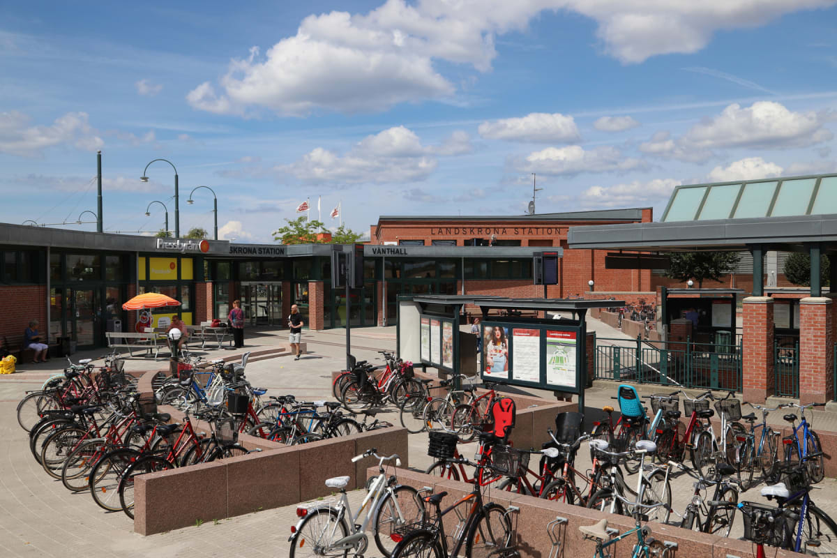 En utomhusbild tagen under dagtid föreställande Landskrona Station med flera cyklar parkerade i förgrunden och stationsbyggnaden i bakgrunden. Stora vita moln syns på en blå himmel och flera personer befinner sig i bildens mitt.