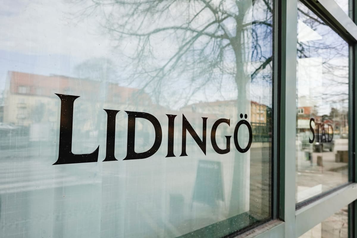 En bild tagen genom en glasruta med ordet "LIDINGÖ" i stora svarta bokstäver. Reflektioner i glaset visar bare trädgrenar, en byggnad med rött tak och en grå himmel. Till höger syns utomhusmiljö och ordet "STAD".