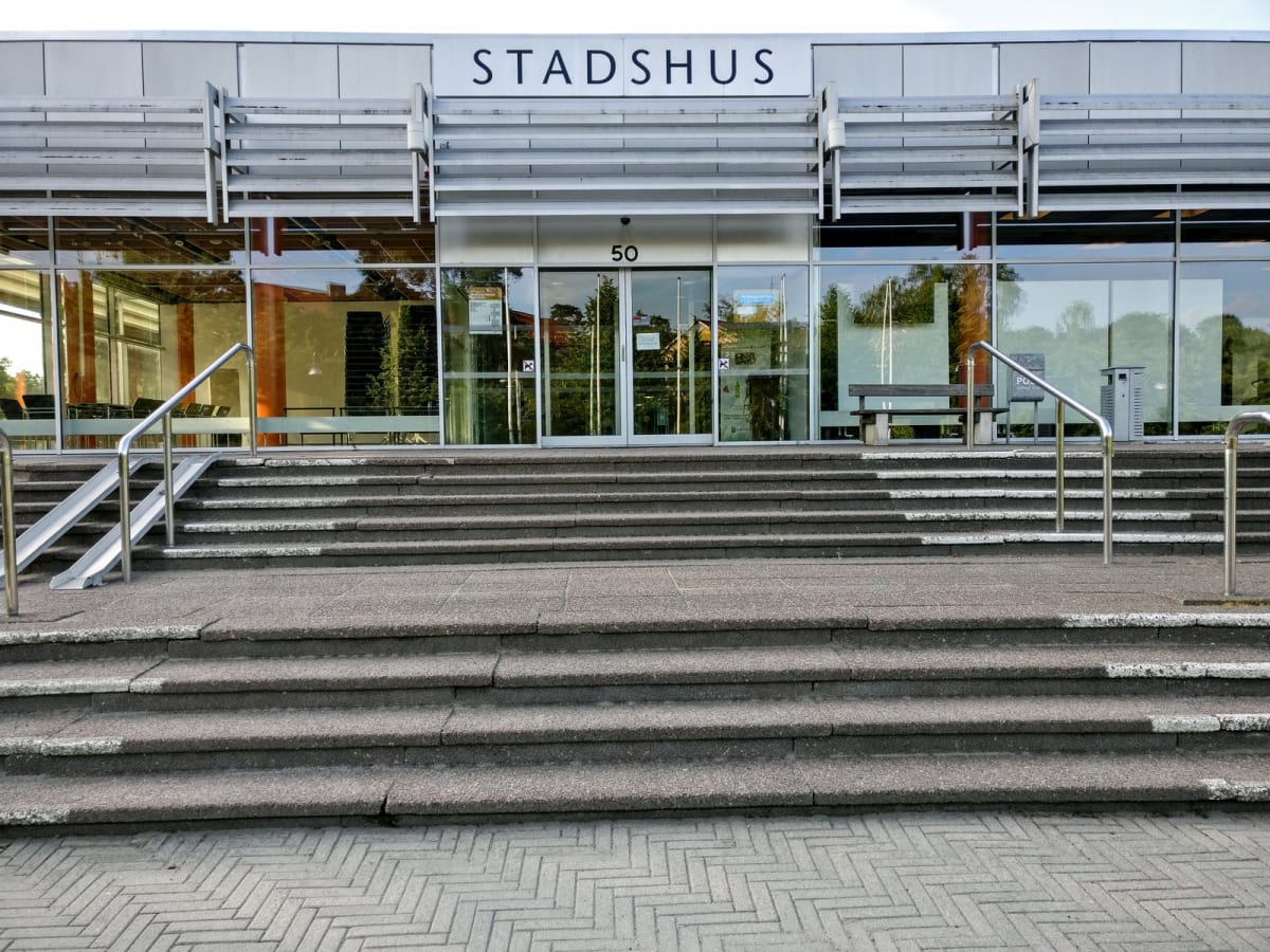 En bild som visar entrén till ett modernt stadshus under dagtid. Ingången har glasdörrar märkta med '50' och texten 'STADSHUS' ovanför. Det finns en bred trappa i betong med ramper och räcken på sidorna, samt en parkbänk och papperskorgar utanför gla