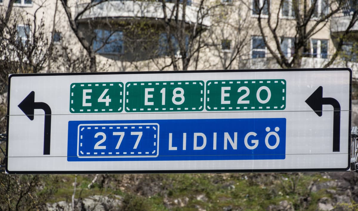 En vägskylt med information om vägar E4, E18, E20 och väg 277 mot Lidingö. Skylten har svarta pilsymboler som visar riktningen för vägarna. I bakgrunden syns delar av en byggnad och träd utan löv.