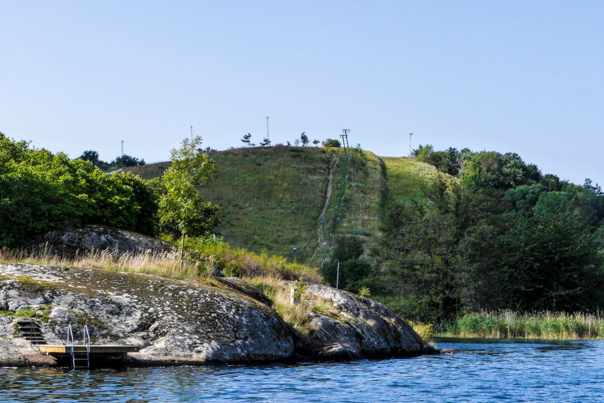 En bild visar ett landskap med en grönskande sluttning där en liftanläggning och en stig syns, samt en klippformation med en brygga och stege ner i vattnet. I förgrunden syns vattenytan.
