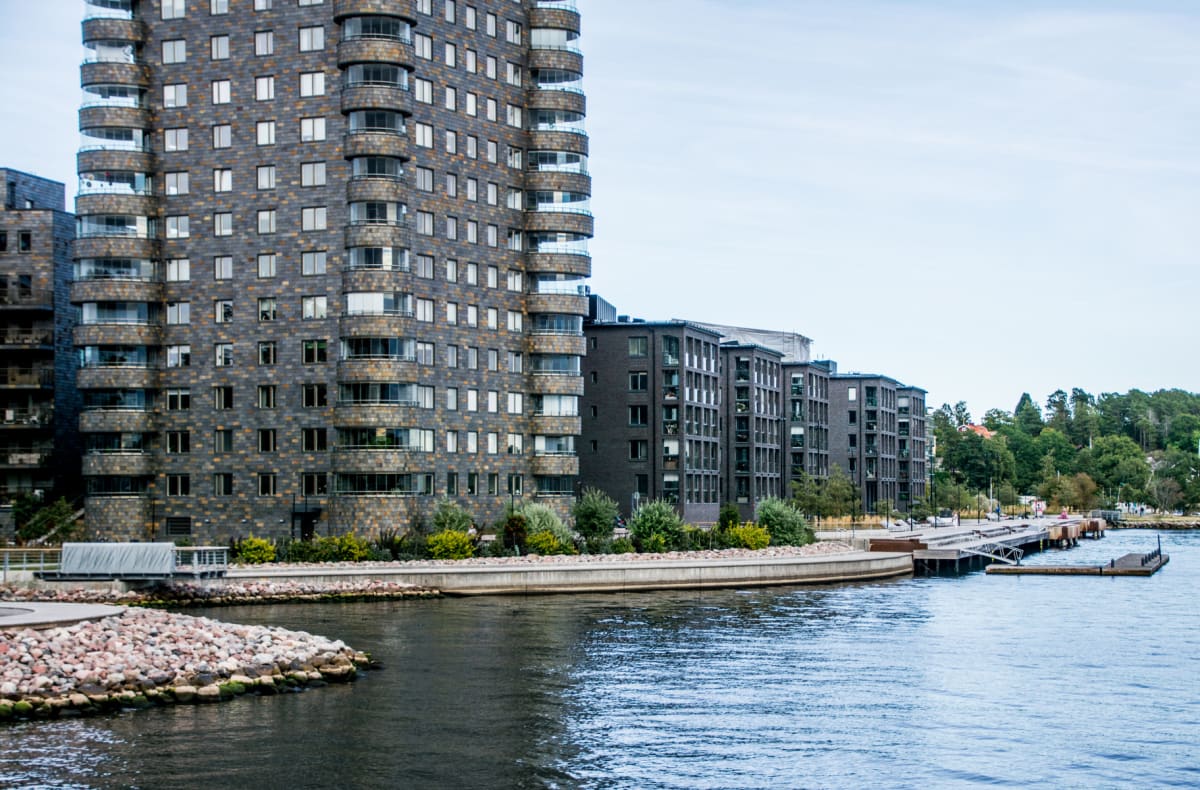 En stor, mörk byggnad med många fönster och balkonger dominerar bilden till vänster. Till höger syns flera mindre, mörka byggnader längs en strandlinje med en brygga och båtplatser. Vattenytan täcker förgrunden och delar av mitten av bilden. En skogs