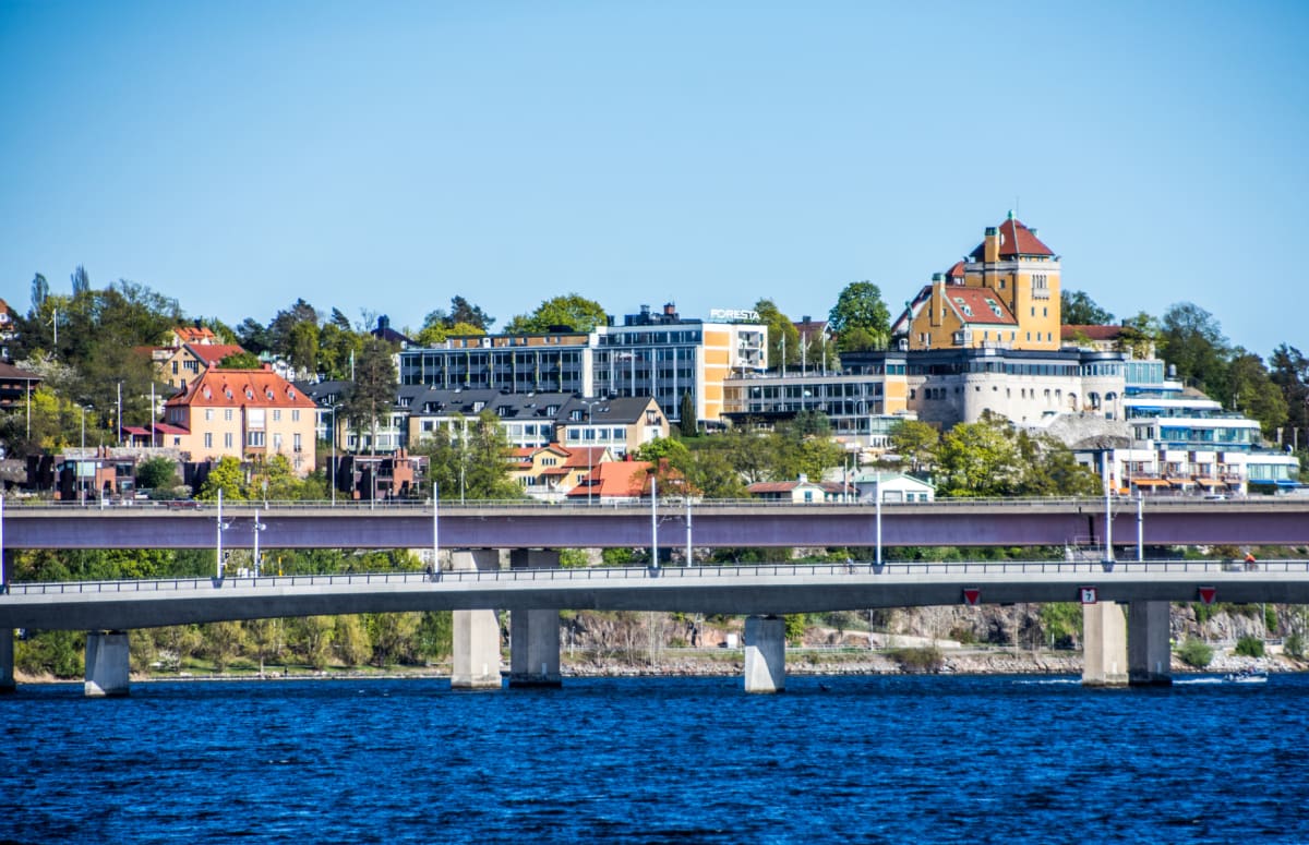 En bild tagen över vatten som visar en bebyggd kustlinje med broar i förgrunden och byggnader på en höjd i bakgrunden. En skylt med texten "FORESTA" är synlig.