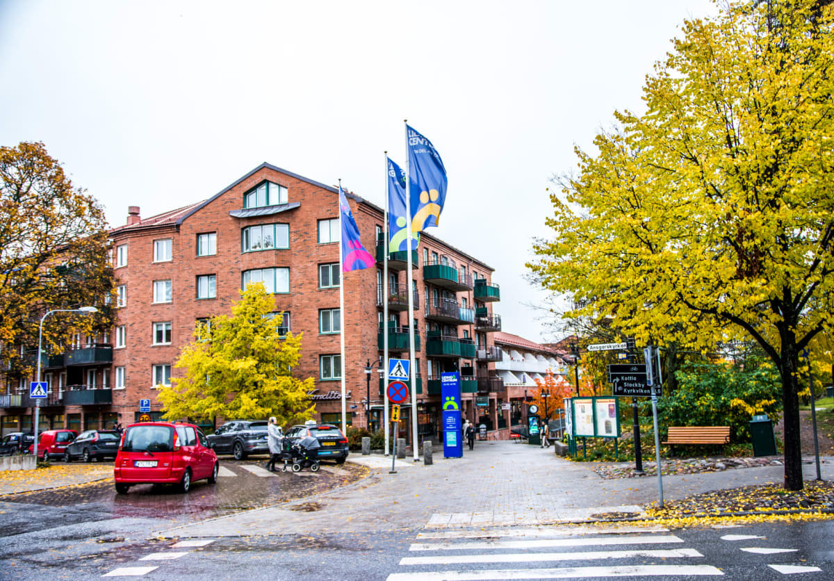En ganska bred bild av en gata i en stad med en röd personbil som vänder mot betraktaren. Till höger finns ett träd med gula löv och till vänster ett tegelhus med balkonger. På gatan syns en person som går med en mörk barnvagn och en bil.