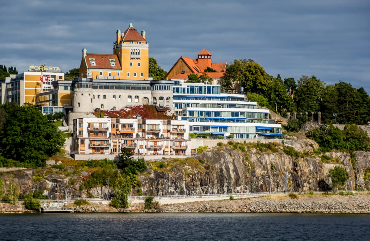 En bild visar en klippig strandkant med mörkblått vatten i förgrunden och en samling komplexa byggnader i olika stilar uppe på klippan. Himmelen är molnig och skog täcker delar av klippan och området bakom byggnaderna.
