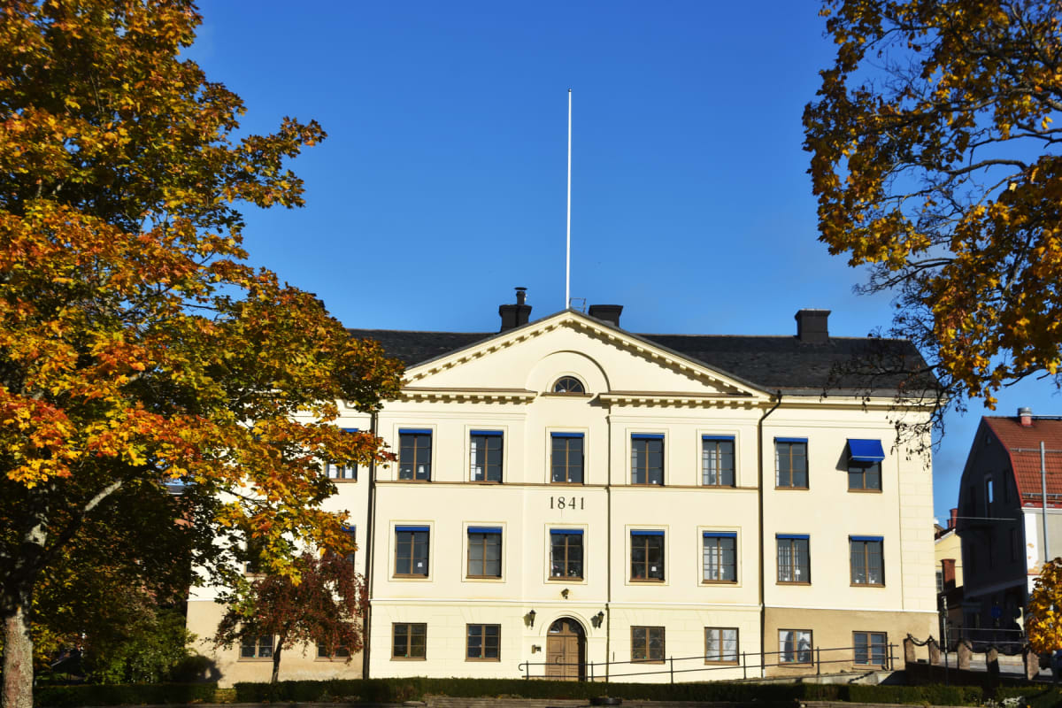 En ljusgul, ståtlig byggnad med mörk tak, blåa fönstermarkiser och texten "1841" centralt placerad. I förgrunden syns höstträd med orangea och gula löv, och en flaggstång reser sig ovanför byggnaden mot en klarblå himmel.