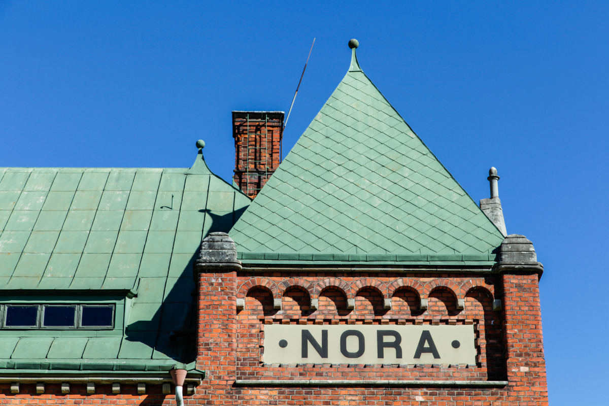 En fasad på en röd tegelbyggnad med grönmålat tak. På tegelfasaden syns ett skylt med texten "NORA" i svart text på ljus bakgrund. Byggnaden har fönster och en skorsten. Hela byggnaden är belyst av solljus med en klarblå himmel i bakgrunden.