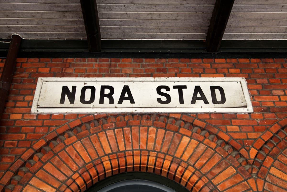 En rektangulär skylt med texten ”NORA STAD” i svarta versaler på vit bakgrund är fäst på en vägg av rött tegel. Under skylten, inbyggd i tegelväggen, finns en rundad valvformad del.