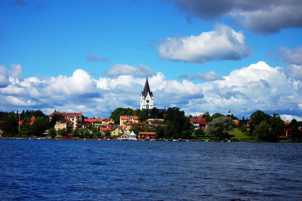 En vidvinkelbild tagen från vattenytan, visar ett samhälle längs en strand med en kyrka med spetsigt torn i centrum mot en blå himmel med moln. Gröna träd och olikfärgade hus med röda tak fyller strandkanten.