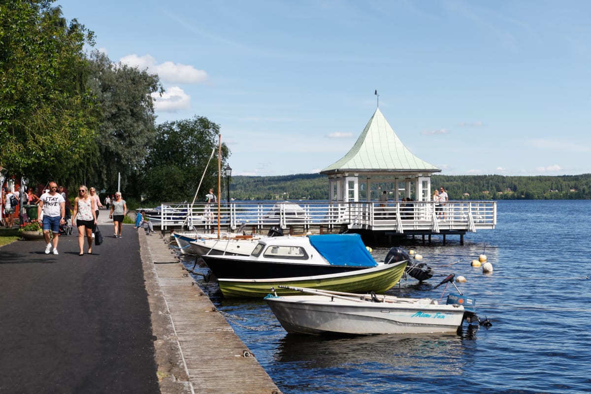 En solig dag vid en vattenkant med en brygga, en sjöpaviljong och flera båtar förtöjda. Människor promenerar längs en gångväg intill vattnet, och i bakgrunden syns skogklädda kullar under en delvis molnig himmel.